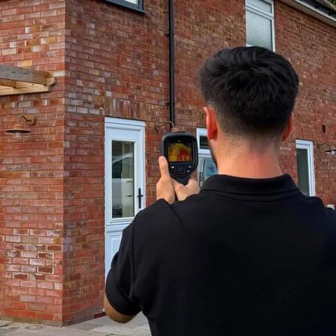 Surveyor using a thermal imaging camera on a red-brick house during a condition survey, homebuyer survey, or building survey.
